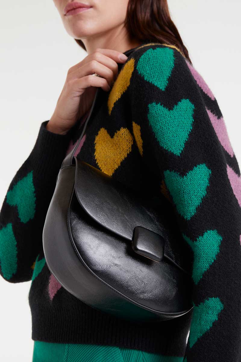 Person wearing a black sweater with colorful heart patterns, holding a black leather bag.
