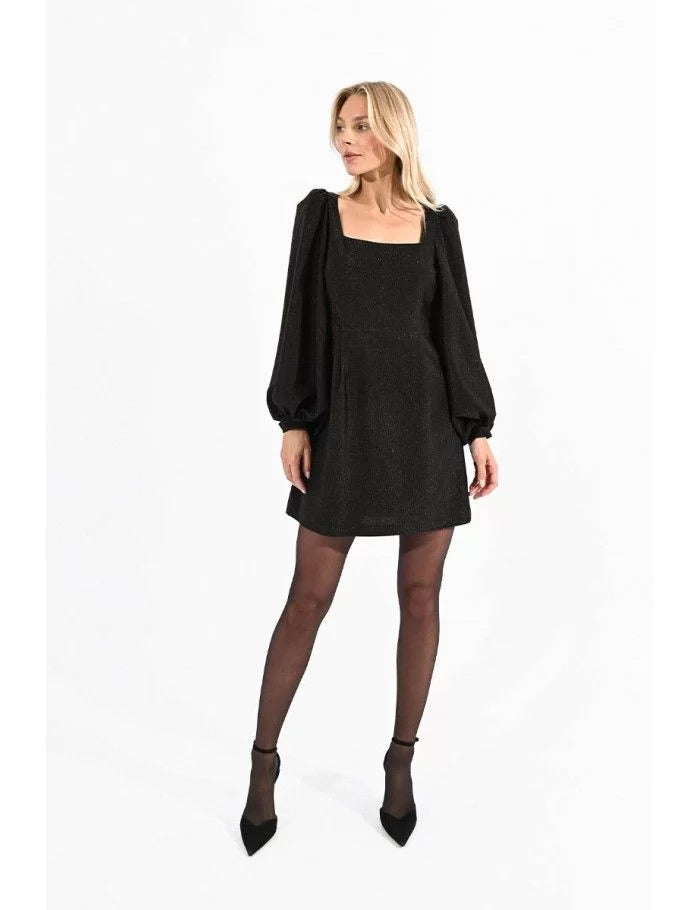 A model wearing a black mini dress with a square neckline and puffed sleeves, paired with sheer tights and black shoes.
